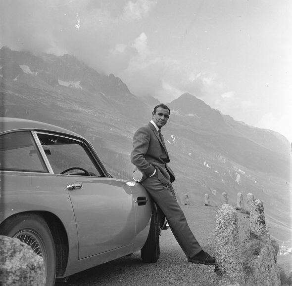 SEAN CONNERY se relaja apoyado sobre su Aston Martin DB5 durante el rodaje de escenas de Goldfinger en los Alpes suizos. Copyright Notice – © 1964 Danjaq, LLC and United Artists Corporation. All rights reserved.