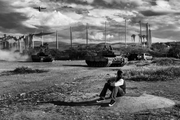 Melilla, Spain April 21st 2014. A young migrant from Mali sits in front of the C.E.T.I while at his back, a couple of tanks of the Spanish army are doing a practice.